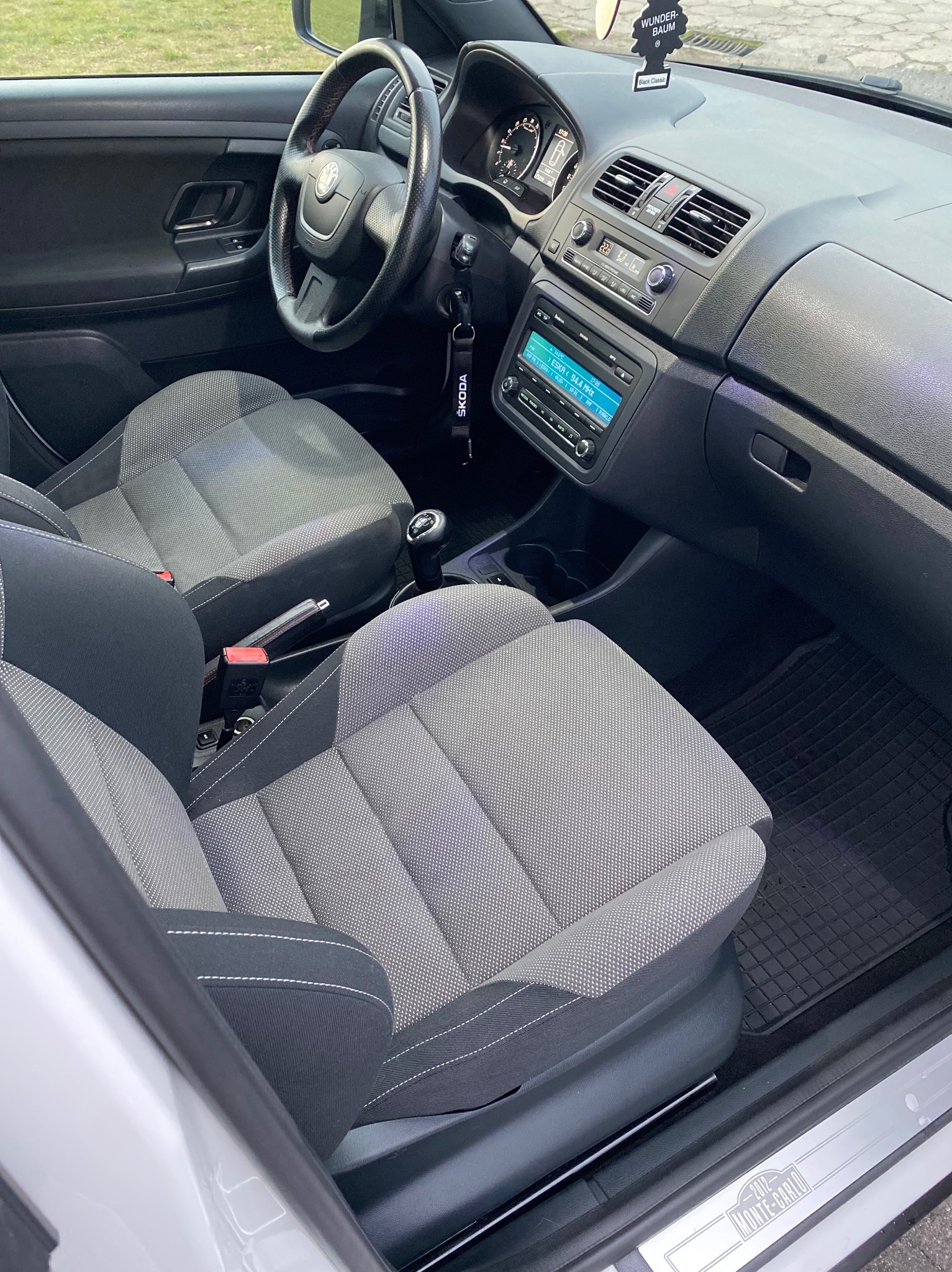Wnętrze samochodu Skoda Fabia Monte Carlo z szaro-czarną tapicerką, widok na fotele, deskę rozdzielczą i kierownicę, z zawieszką zapachową Wunder-Baum. Widoczny napis Monte Carlo na progu.