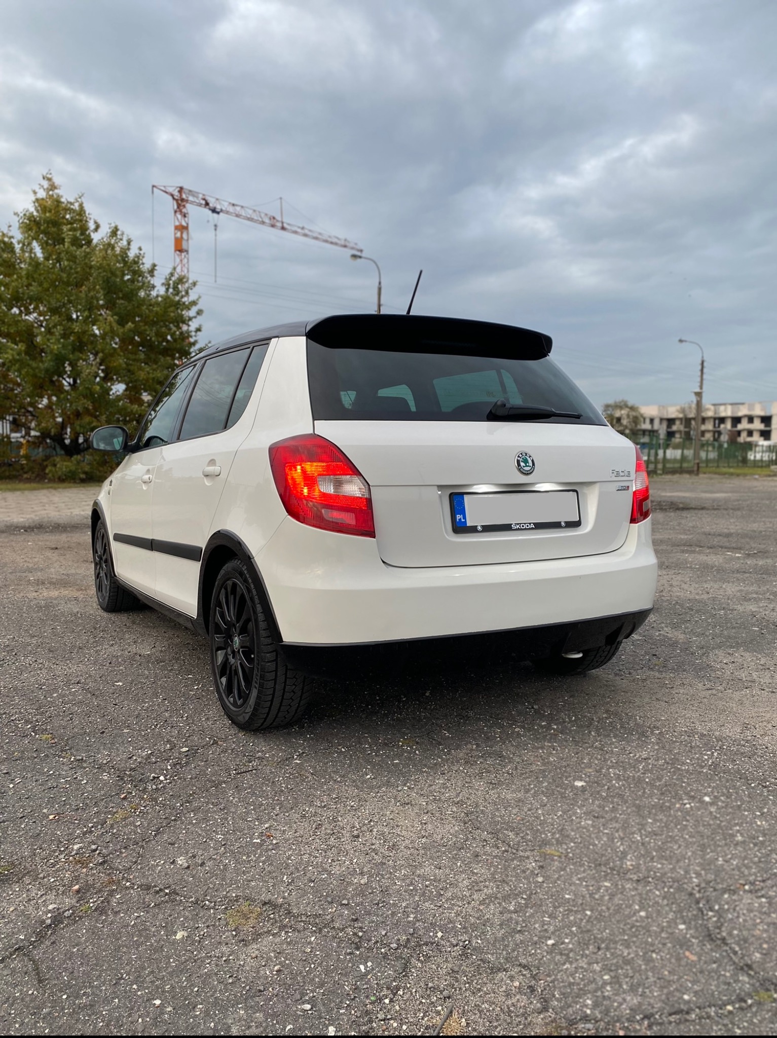 Biała Skoda Fabia z czarnym dachem i felgami, widziana od tyłu na betonowym placu z budową w tle i pochmurnym niebem.