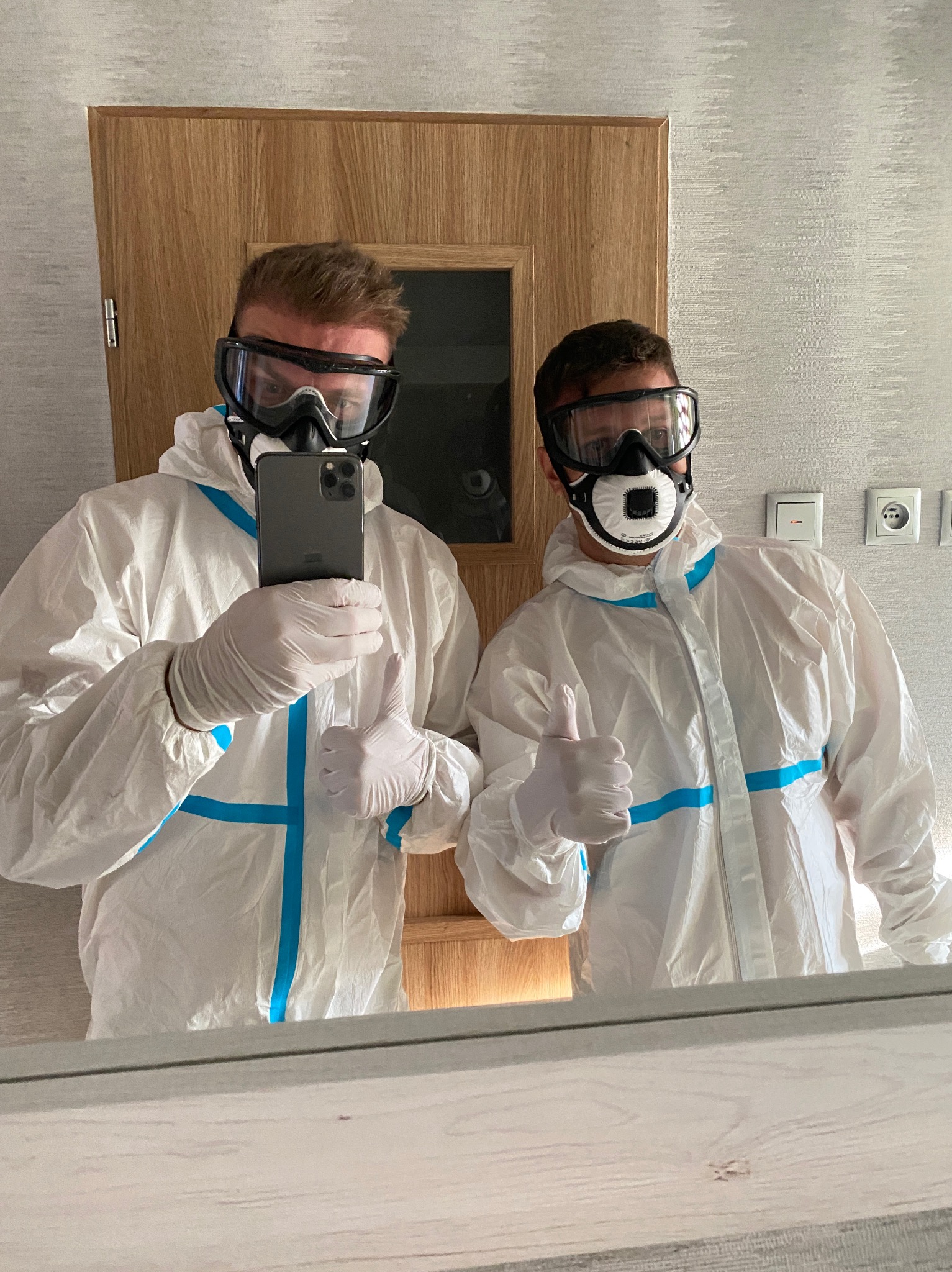 Dwaj mężczyźni w białych kombinezonach ochronnych z niebieskimi paskami i maskach ochronnych robią sobie selfie, pokazując kciuki w górę w pomieszczeniu z drewnianymi drzwiami i szarą tapetą.