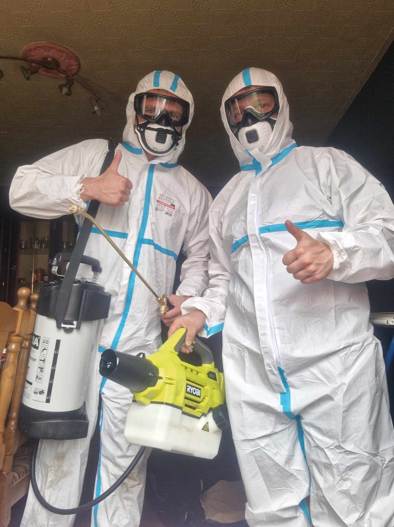 Dwóch pracowników w białych kombinezonach ochronnych i maskach, jeden z opryskiwaczem plecakowym, drugi z elektrycznym zamgławiaczem Ryobi, obaj z podniesionymi kciukami.