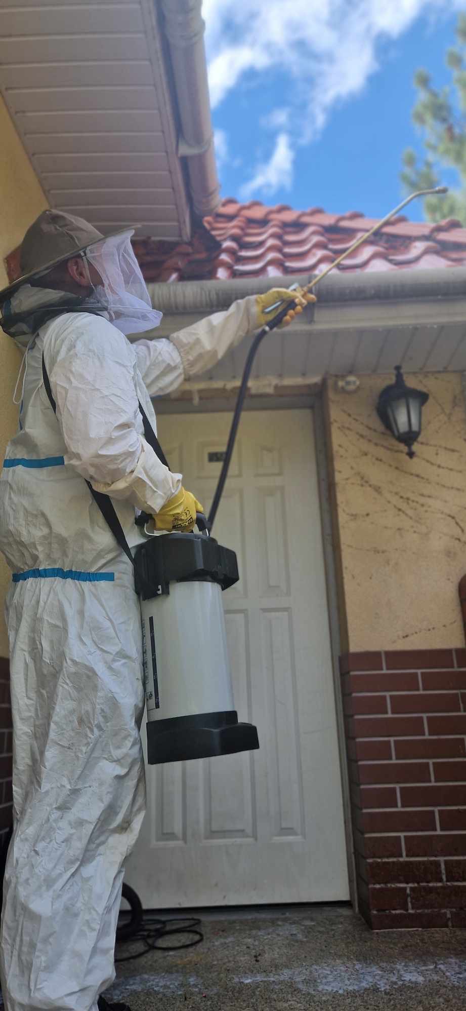 Pracownik w kombinezonie ochronnym, siatce na twarzy i żółtych rękawicach spryskuje opryskiwaczem okolice dachu domu z czerwonej dachówki i białych drzwi wejściowych.
