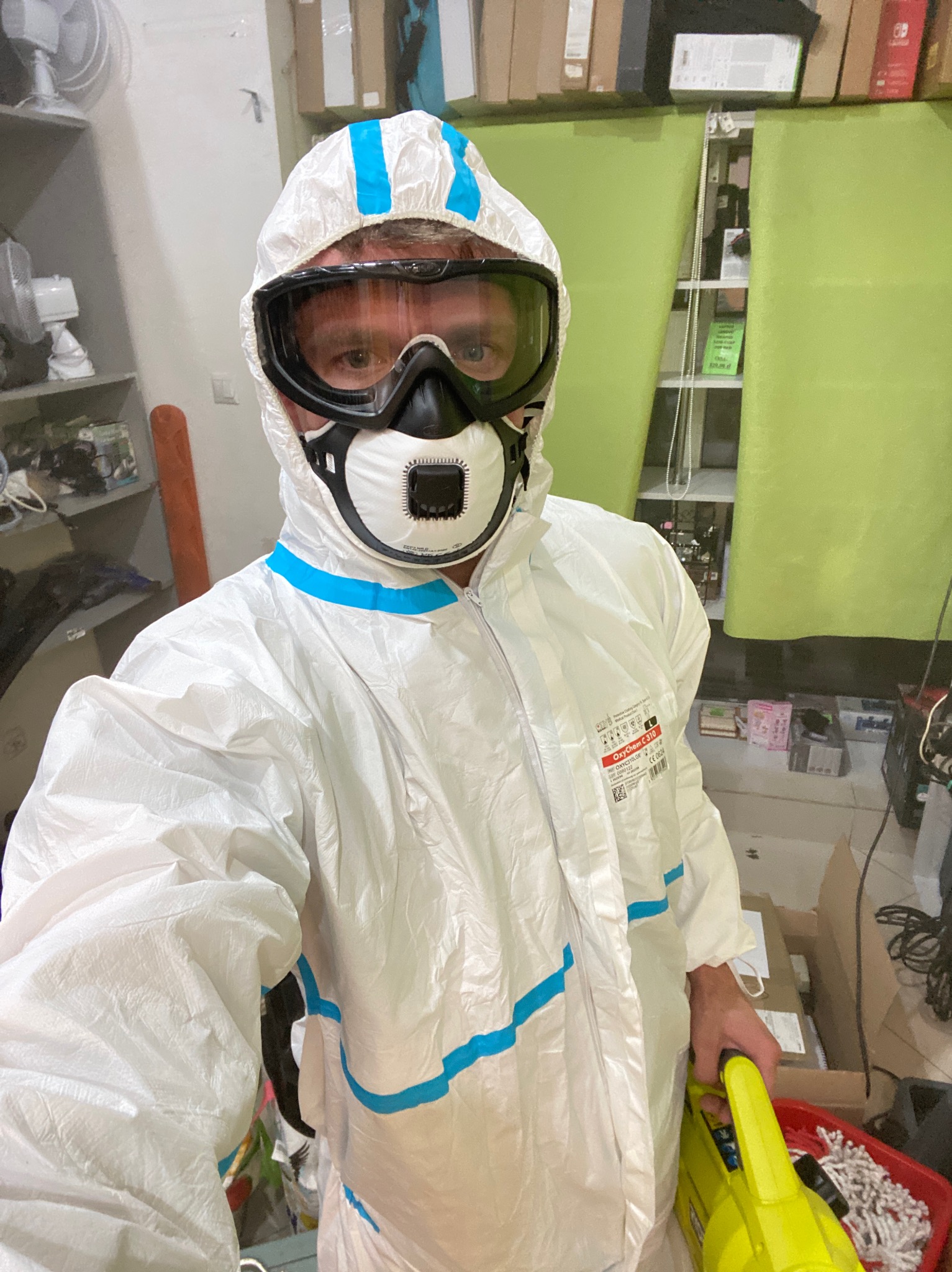 Osoba w kombinezonie ochronnym, masce i goglach, trzymająca żółtą lampę roboczą na tle półek z różnymi przedmiotami.