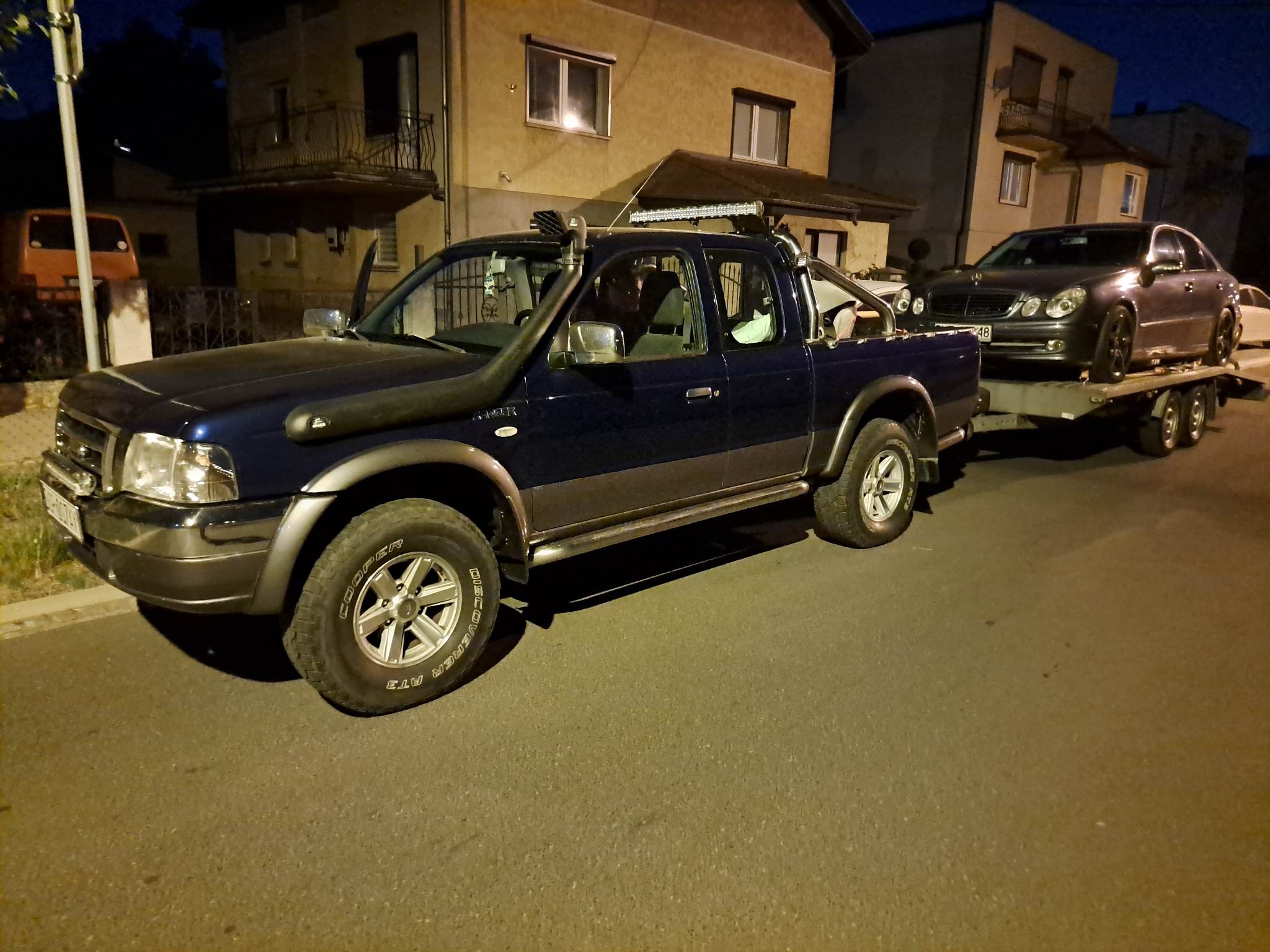 Niebieski pickup z orurowaniem i światłami LED ciągnie lawetę z szarym sedanem na tle budynków mieszkalnych. Nocne oświetlenie uliczne.