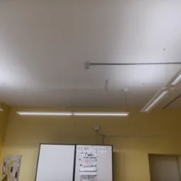 Wnętrze klasy z oświetleniem LED, czujnikiem dymu na suficie i tablicą z notatkami.