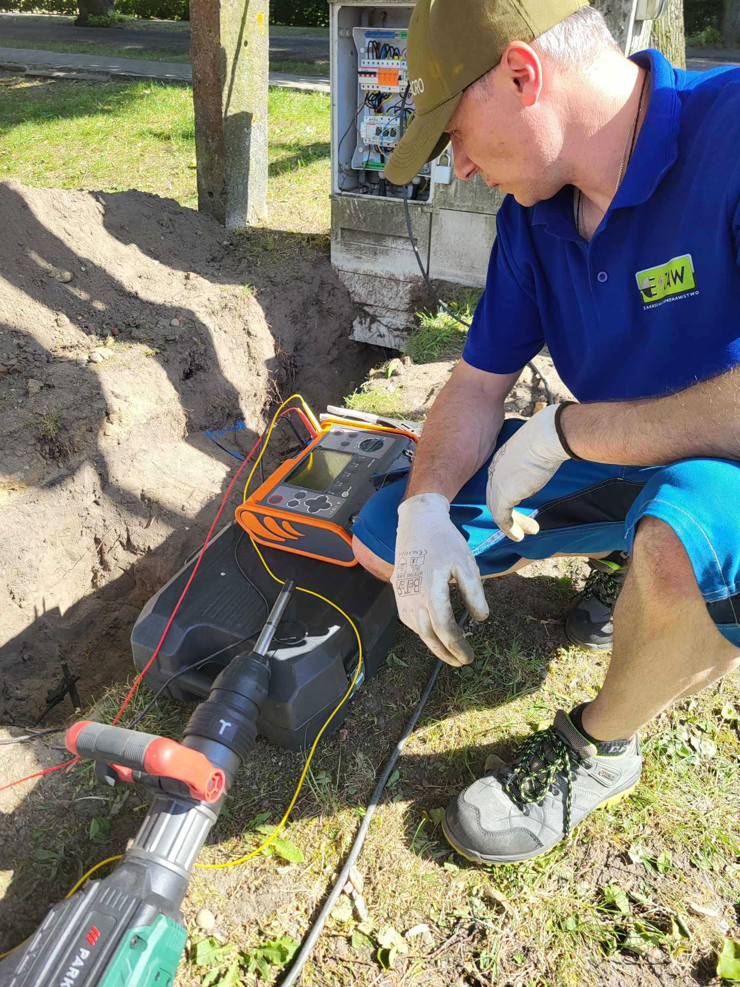 Elektryk w niebieskiej koszulce polo i czapce z daszkiem, w trakcie pomiarów elektrycznych przy otwartej skrzynce elektrycznej na zewnątrz, używa miernika i wiertarki udarowej, obok wykopany dół.
