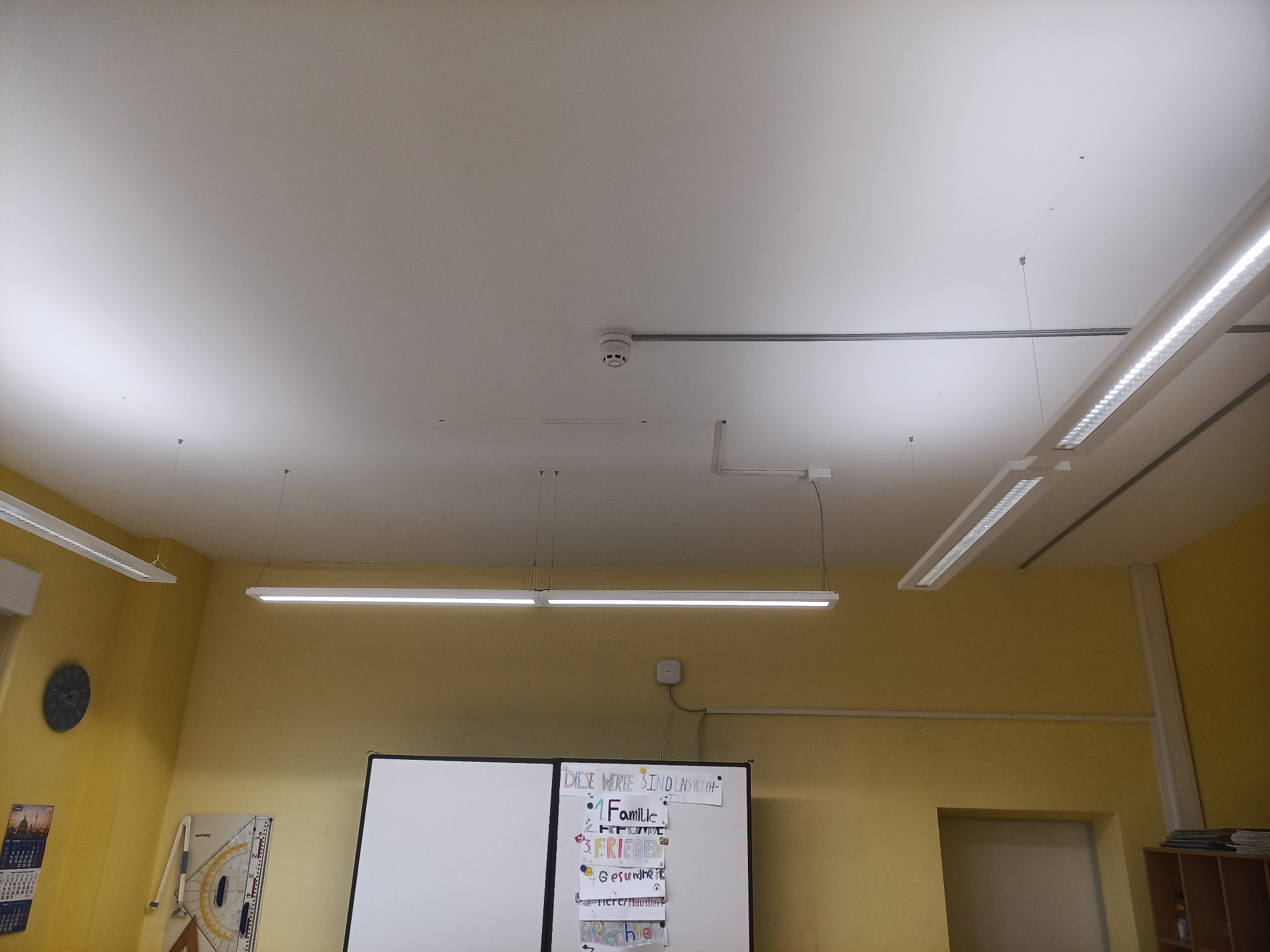 Wnętrze klasy z oświetleniem LED, czujnikiem dymu na suficie i tablicą z notatkami.