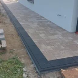 Ułożona ścieżka z kostki brukowej w dw&oacute;ch kolorach, brązowo-beżowym i czarnym, prowadząca wzdłuż ściany domu z widocznymi wystającymi przewodami elektrycznymi i oknem.