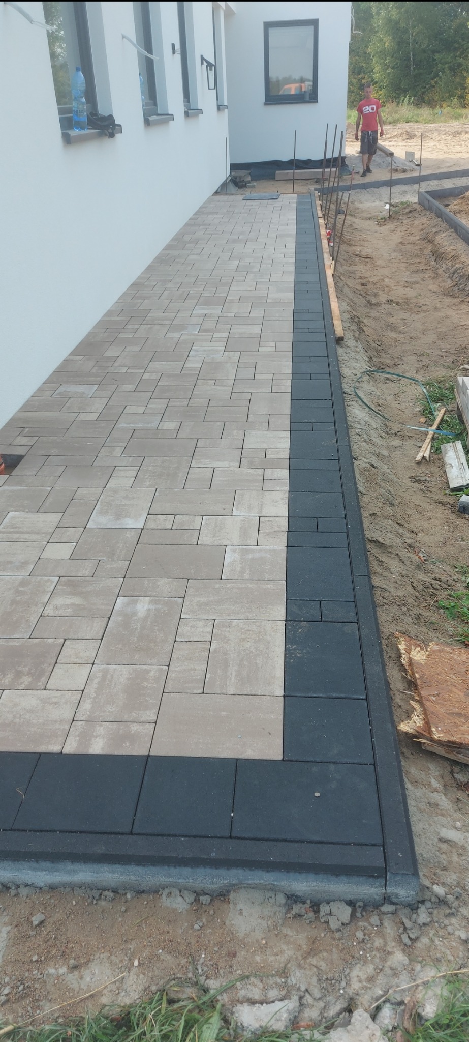 Ułożona ścieżka z kostki brukowej w odcieniach beżu i szarości, obramowana ciemną kostką, przylegająca do białej ściany budynku, z widocznym fragmentem terenu w trakcie prac wykończeniowych.