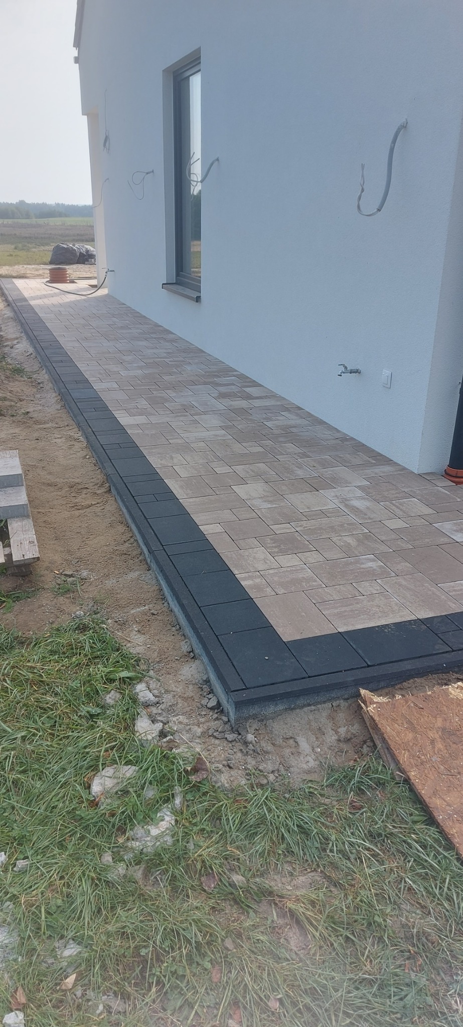 Ułożona ścieżka z kostki brukowej w dwóch kolorach, brązowo-beżowym i czarnym, prowadząca wzdłuż ściany domu z widocznymi wystającymi przewodami elektrycznymi i oknem.