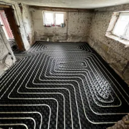 Białe rury ogrzewania podłogowego ułożone w pętle na czarnej izolacji w surowym pomieszczeniu z ceglanymi ścianami i dwoma oknami.