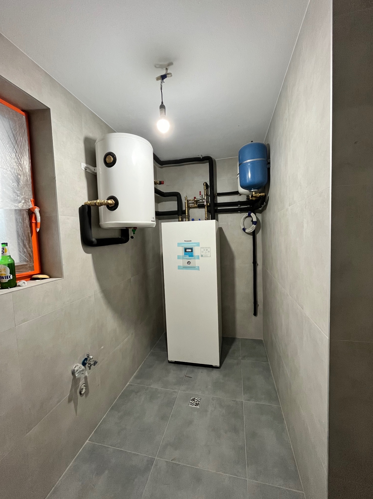 Wnętrze kotłowni z zamontowanym białym zasobnikiem ciepłej wody, niebieskim zbiornikiem wyrównawczym i pompą ciepła Panasonic Aquarea, widoczne rury instalacji grzewczej i wodnej.