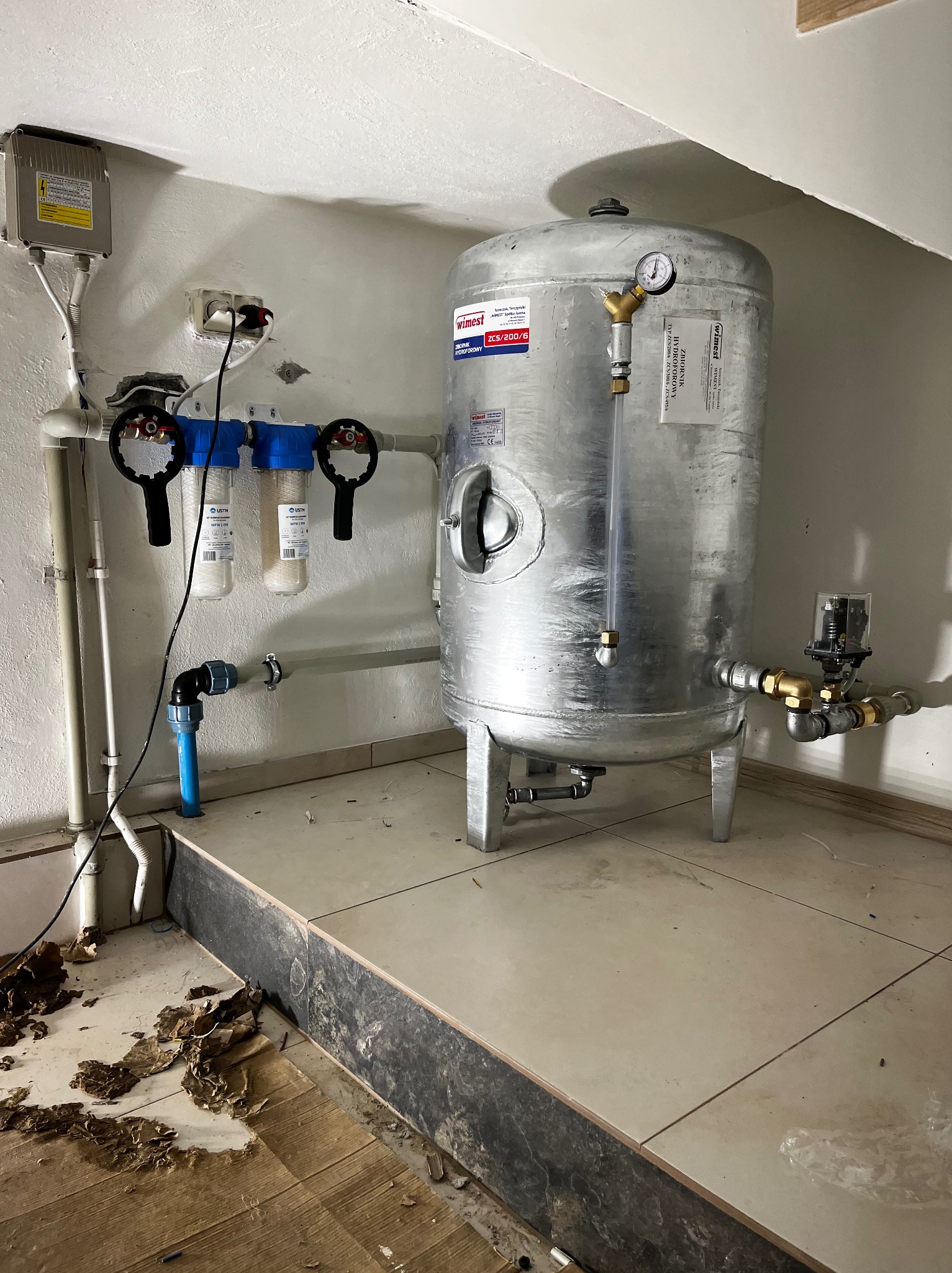 Hydrofor ze srebrnym zbiornikiem ciśnieniowym Wilmest ZCS/200/6, manometrem i filtrami wody, zainstalowany w pomieszczeniu technicznym z widocznymi śladami prac wykończeniowych na podłodze.