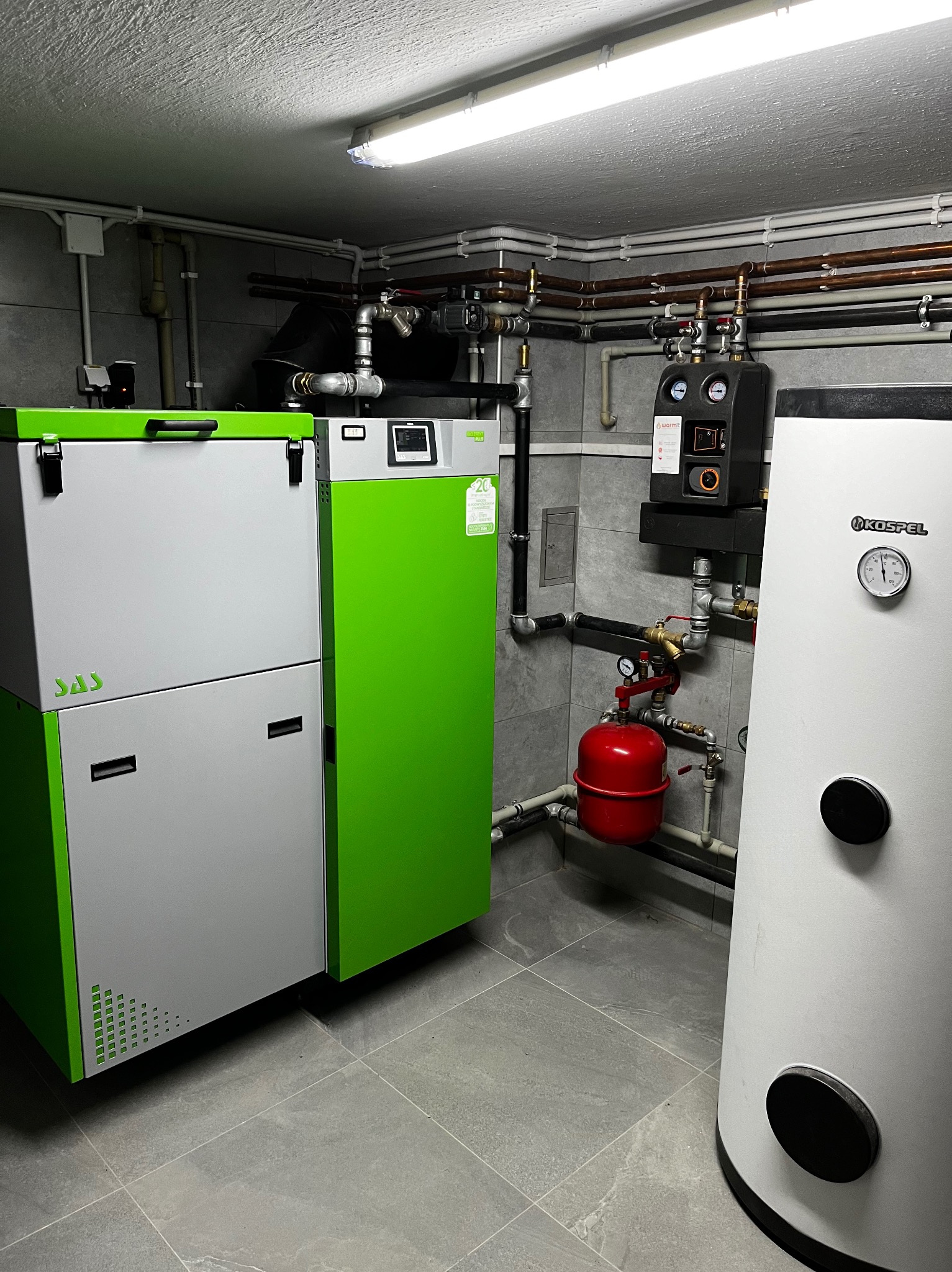 Kotłownia z nowoczesnym piecem SAS w kolorze zielono-szarym, zasobnikiem Kospel z termometrem, czerwonym naczyniem przeponowym i rozbudowaną instalacją z miedzianych rur.