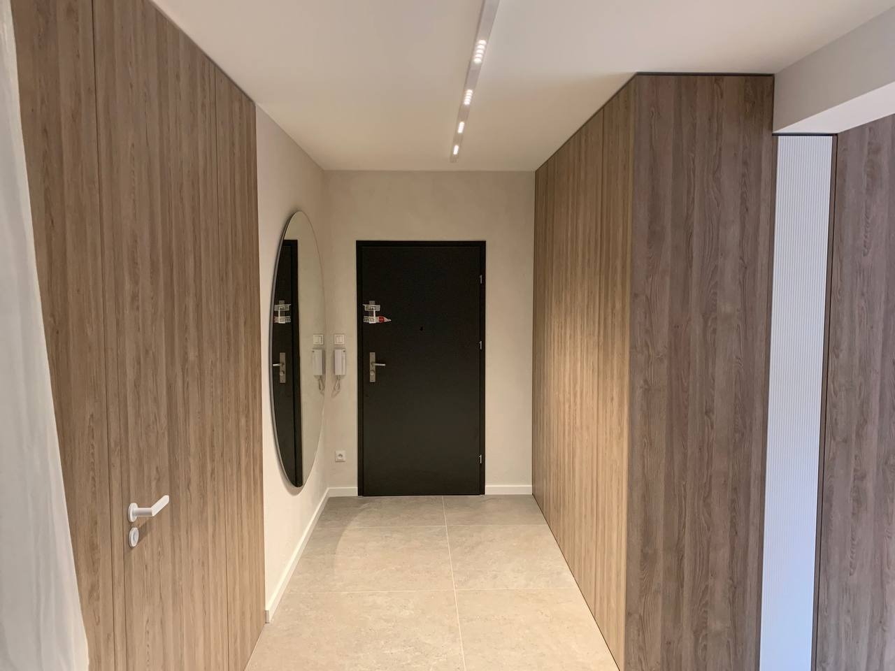 Nowoczesny przedpokój z zabudową drewnopodobną, okrągłym lustrem i czarnymi drzwiami wejściowymi.