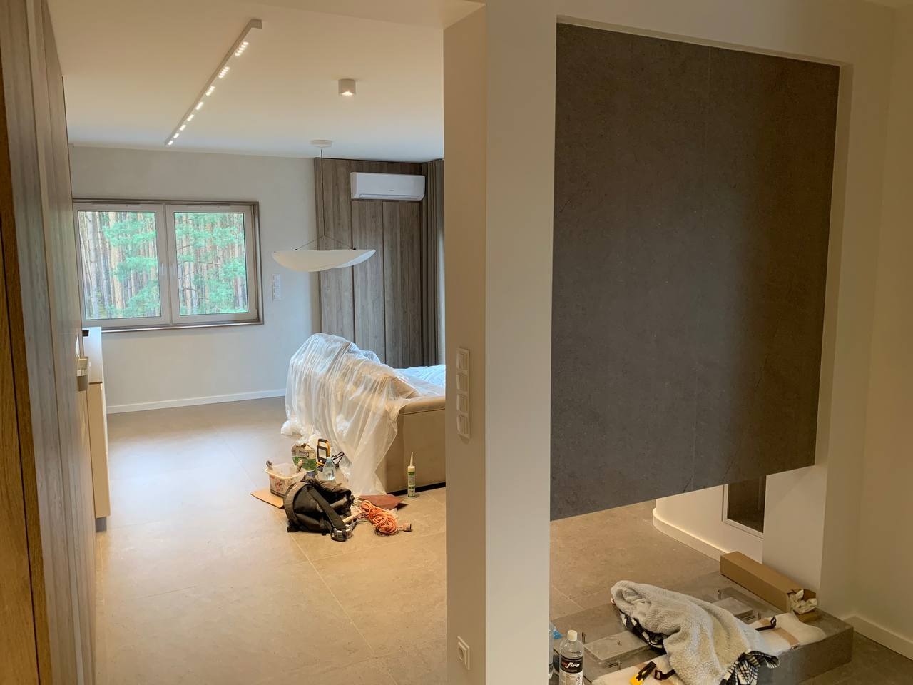 Wnętrze w trakcie prac wykończeniowych: widoczna ściana wyłożona szarymi płytami imitującymi kamień, zabezpieczony folią mebel oraz narzędzia i materiały budowlane na podłodze z płytek.