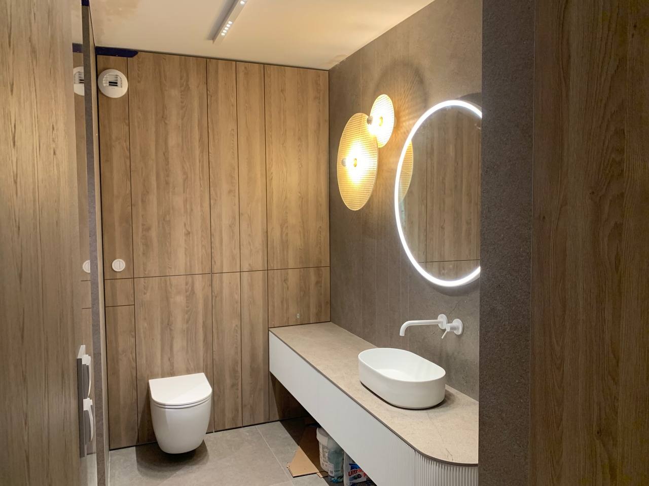Nowoczesna łazienka z drewnopodobnymi panelami ściennymi, okrągłym lustrem z podświetleniem LED, owalną umywalką nablatową i wiszącą toaletą.