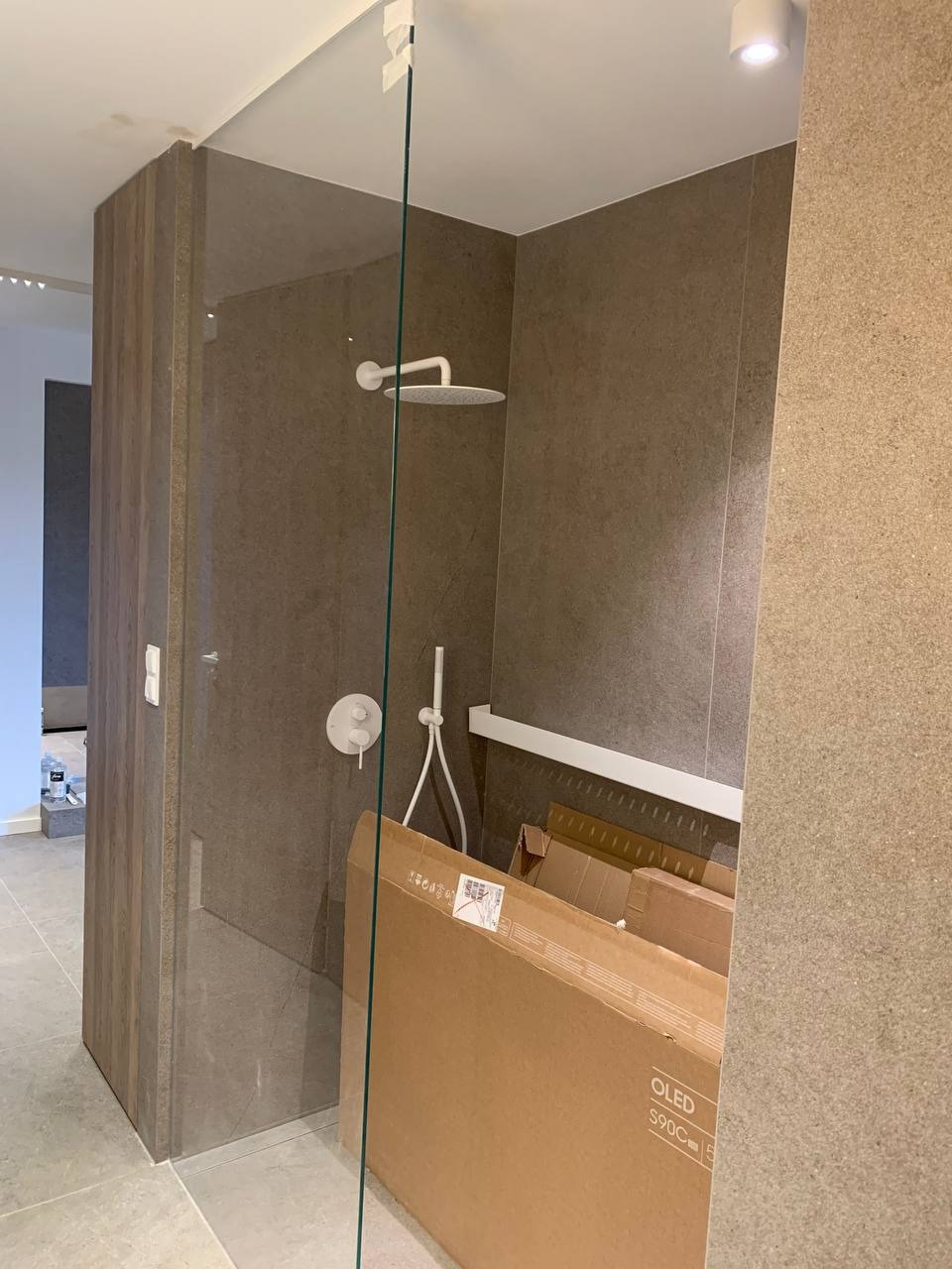 Szklana kabina prysznicowa z zamontowaną deszczownicą i baterią podtynkową, widoczne kartony po sprzęcie RTV, szare płytki na ścianach i podłodze.