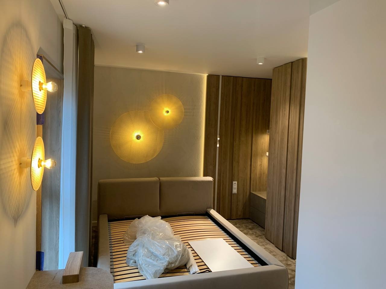 Sypialnia w trakcie urządzania: drewniane panele ścienne, dekoracyjne lampy w kształcie ażurowych kół, łóżko bez materaca z folią ochronną, surowa ściana w tle.