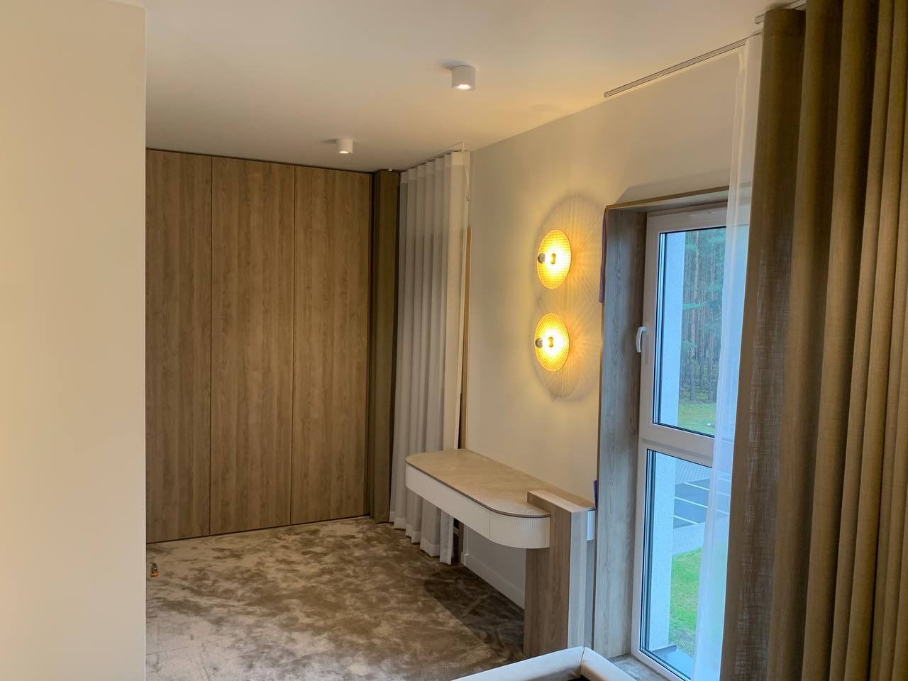 Jasne wnętrze pokoju z zabudową szafy z drewnopodobnymi frontami, toaletką przy oknie z ozdobnymi lampami i zasłonami w odcieniach beżu.