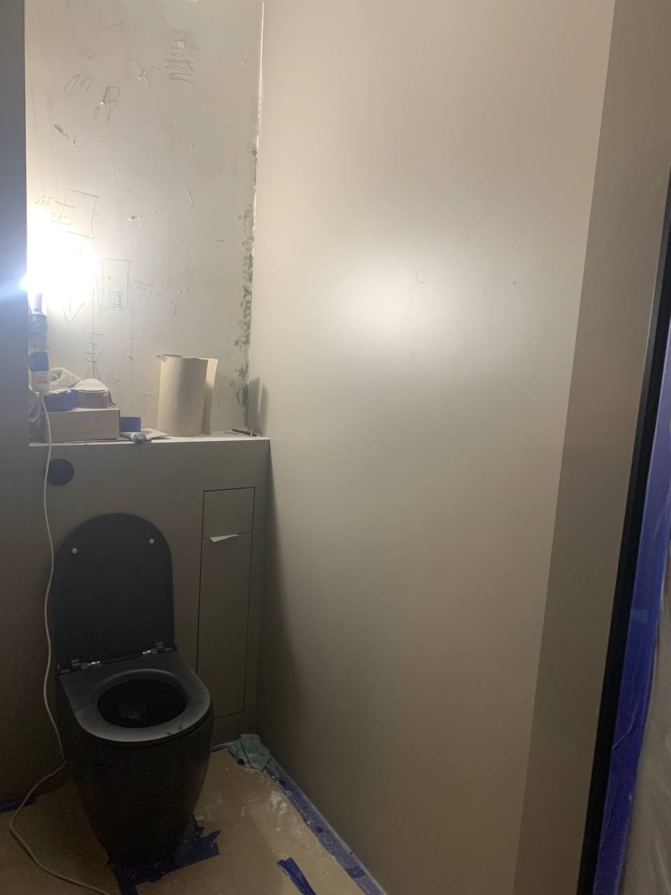 Ciemna toaleta kompaktowa w trakcie montażu w małej łazience, ściany przygotowane do malowania, widoczne ślady po wcześniejszych pracach, zabezpieczona folią podłoga.