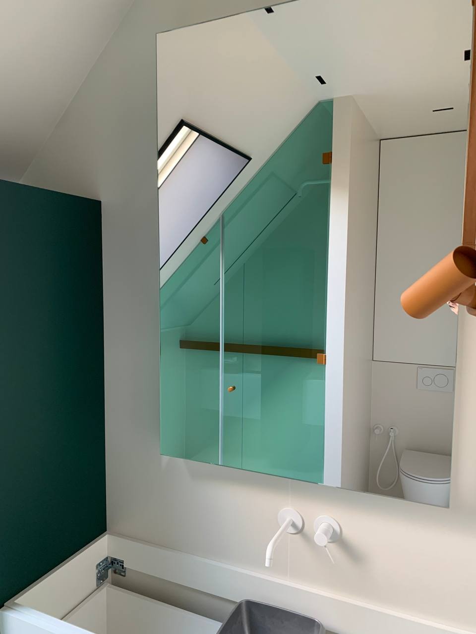 Nowoczesna łazienka z zielonymi akcentami, widoczna w lustrze kabina prysznicowa ze skośnym sufitem i oknem dachowym, minimalistyczna umywalka z białymi bateriami.