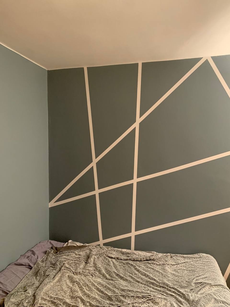 Szara ściana z geometrycznym wzorem z jasnych pasów nad łóżkiem z szarą pościelą.