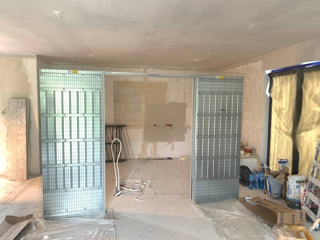 Montaż metalowej konstrukcji do ścian działowych z płyty karton-gips w surowym wnętrzu, widoczne instalacje elektryczne i materiały budowlane na podłodze.