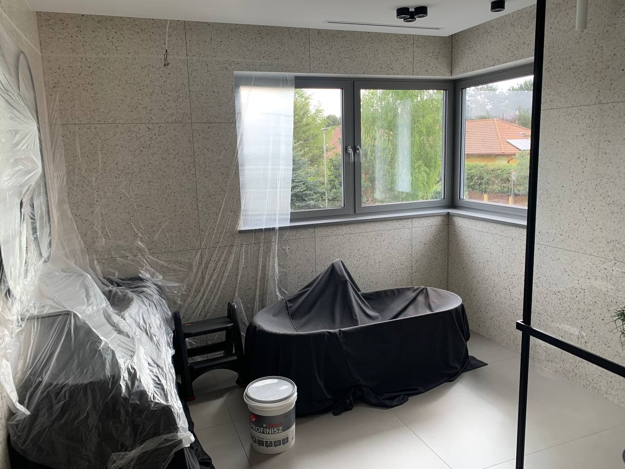 Łazienka w trakcie prac zabezpieczających: wanna zakryta czarną folią, ściany wyłożone płytkami imitującymi lastryko, okno z szarą ramą, wiadro z farbą 'Profinisz' na podłodze.