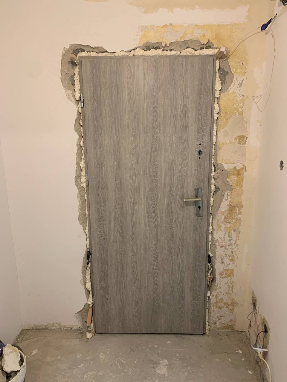 Montaż szarych drzwi wewnętrznych z ościeżnicą, pianka montażowa widoczna wokół ramy, ściany w trakcie przygotowania do wykończenia.