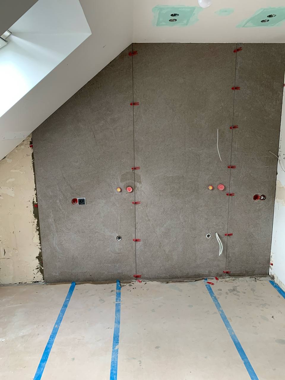 Ściana w trakcie układania szarych płytek imitujących kamień, z czerwonymi klipsami poziomującymi, widoczne gniazdka elektryczne i kable, podłoga zabezpieczona niebieską taśmą malarską, pokój...