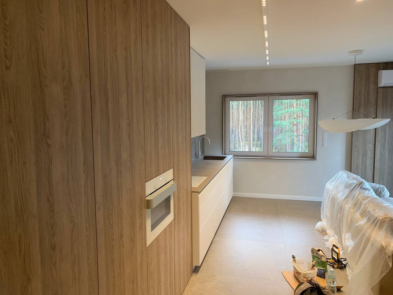 Nowoczesna kuchnia z drewnianymi frontami szafek, wbudowanym piekarnikiem i jasnym blatem, okno z widokiem na las, podłoga w odcieniach szarości, elementy zabezpieczone folią ochronną.