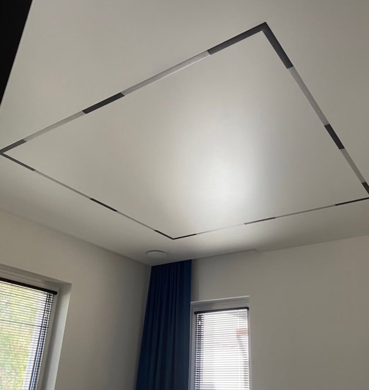 Biały sufit z kwadratowym, podświetlanym obramowaniem, widok z dołu. W tle okna z żaluzjami i granatowa zasłona.