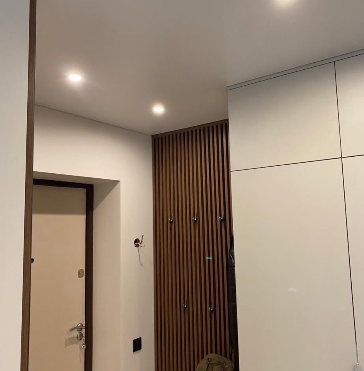 Nowoczesny przedpokój z białymi szafami, drewnianą ścianką z haczykami i podwieszanym oświetleniem punktowym. Widoczne drzwi wejściowe.