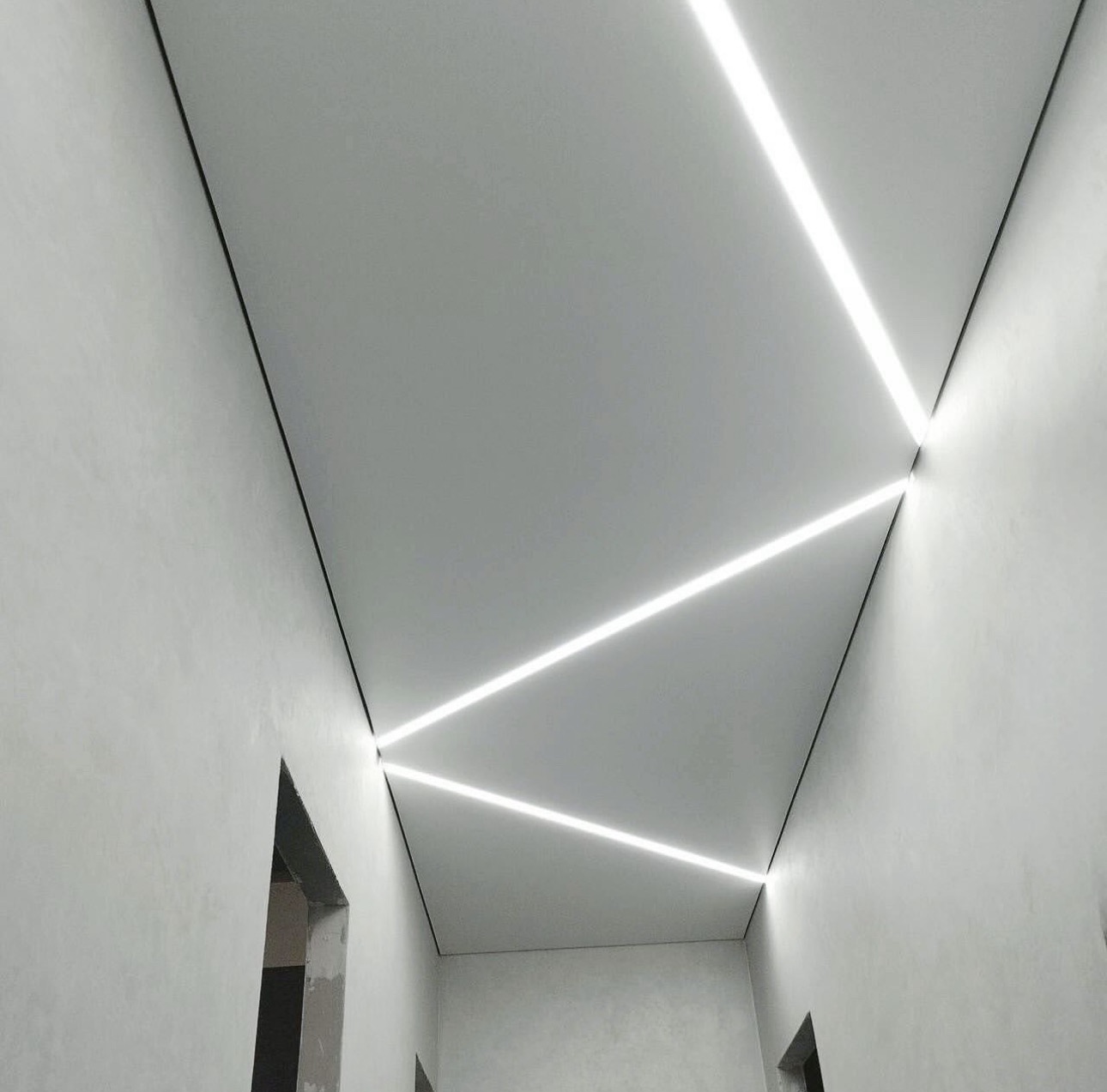 Nowoczesny sufit z wbudowanym oświetleniem LED tworzącym geometryczny wzór. Minimalistyczne wykończenie ścian i widoczne fragmenty otworów drzwiowych.