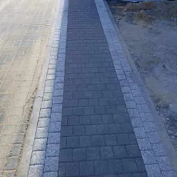 Ścieżka z ciemnej kostki brukowej obramowana jasną kostką granitową, prowadząca wzdłuż ściany budynku, z widocznym piaskiem i ziemią po bokach.