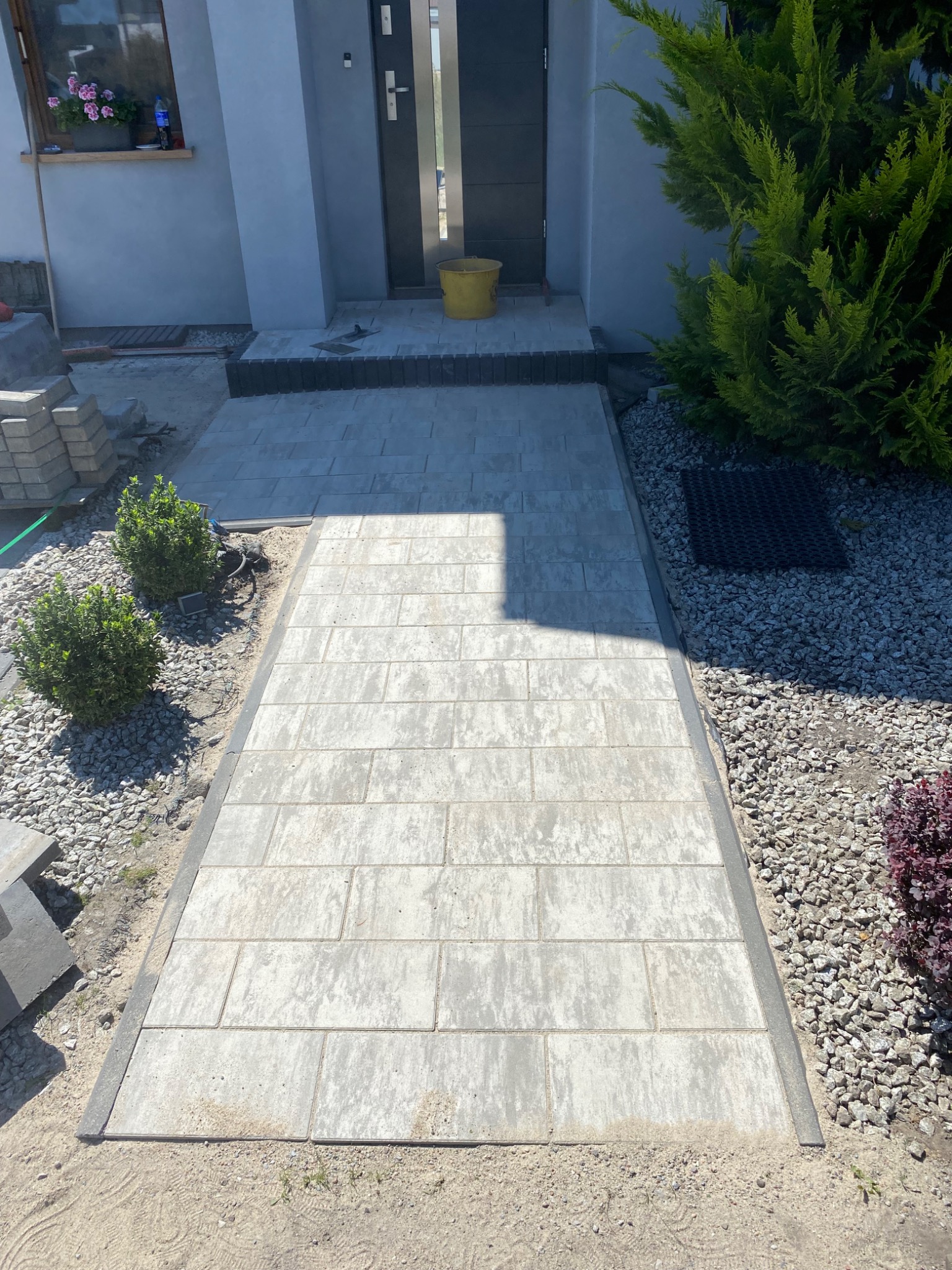 Ułożona ścieżka z kostki brukowej prowadząca do domu, obrzeża z ciemniejszej kostki, po bokach ozdobne krzewy i wysypane kamyki.