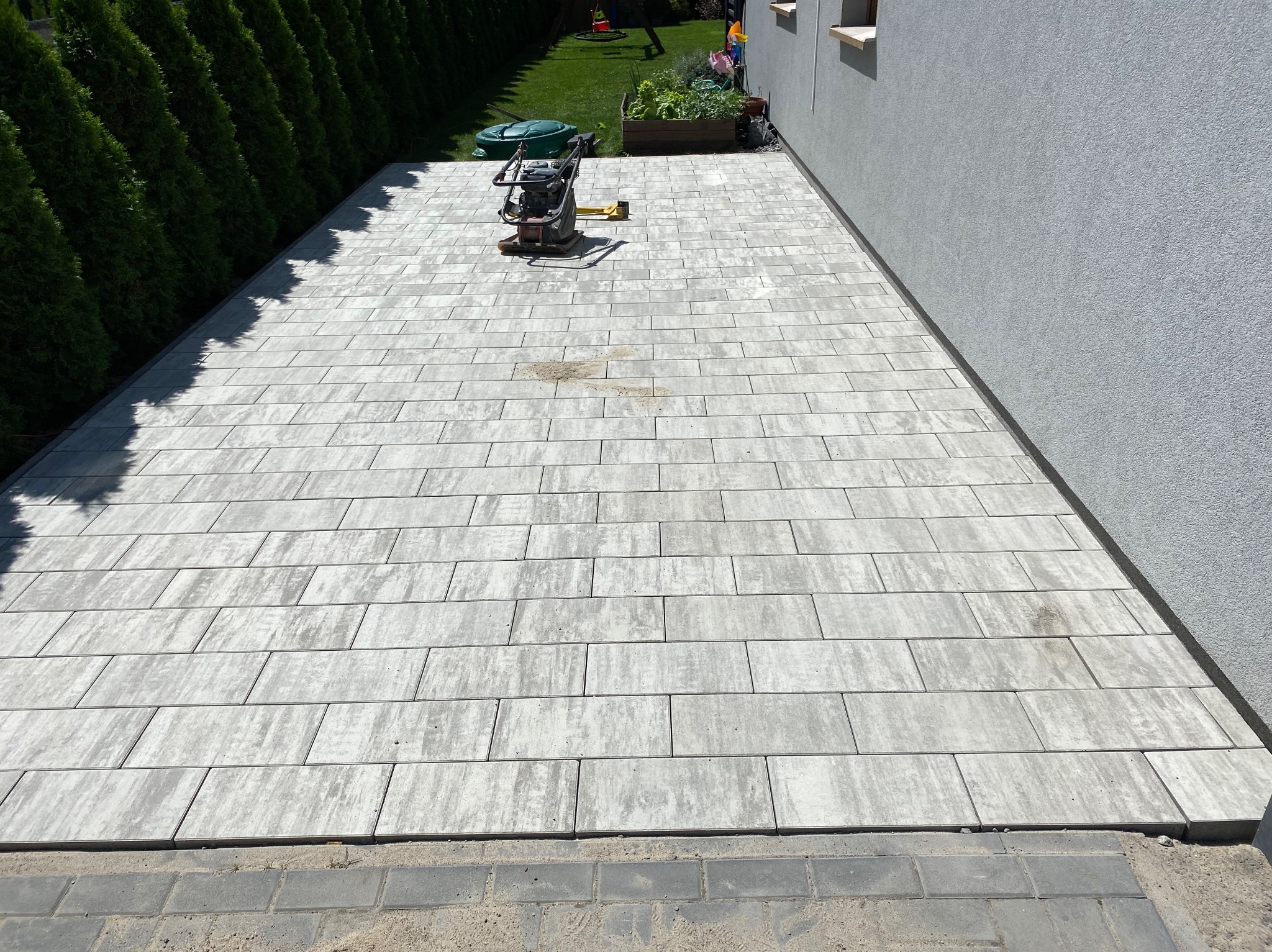 Świeżo ułożona, szara kostka brukowa o prostokątnym kształcie, częściowo ubita zagęszczarką mechaniczną, widok z góry na tle zielonego trawnika i elewacji budynku.