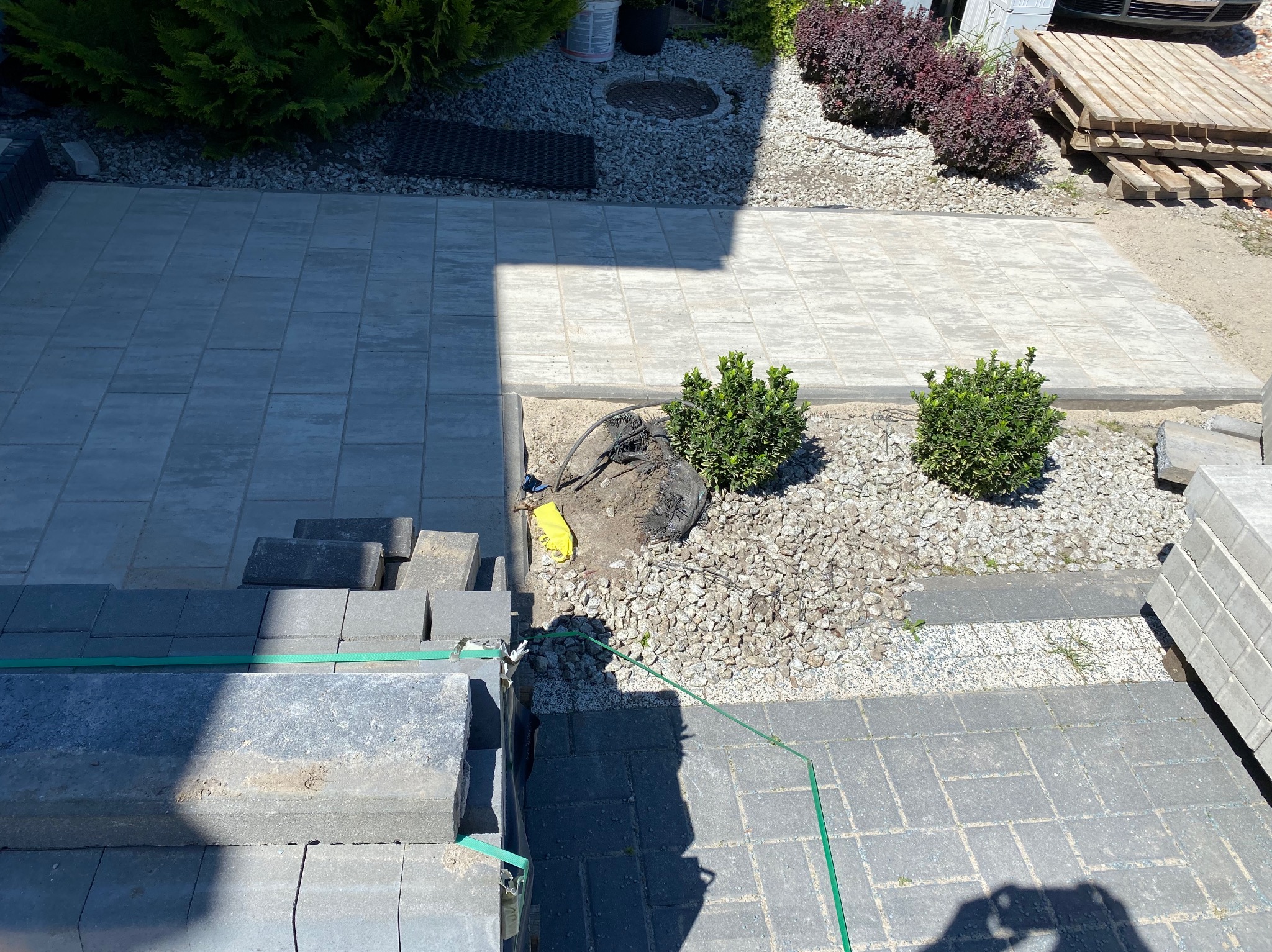 Ułożona szara kostka brukowa na posesji, z widocznymi paletami z kostką, roślinnością i fragmentem węża ogrodowego, sugerująca trwające prace wykończeniowe.