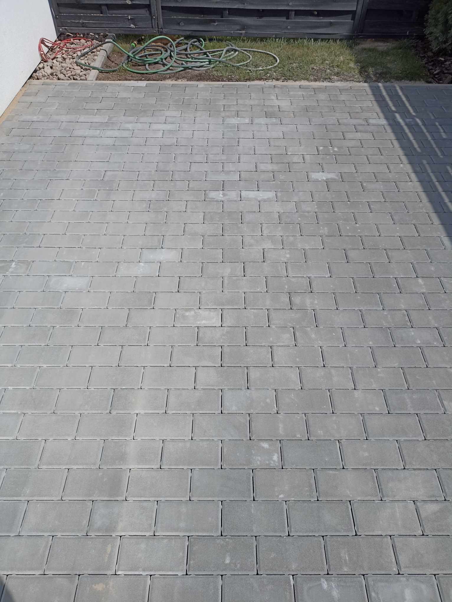 Ułożona szara kostka brukowa na podwórku, widoczny fragment trawnika z wężem ogrodowym i drewnianym płotem w tle.