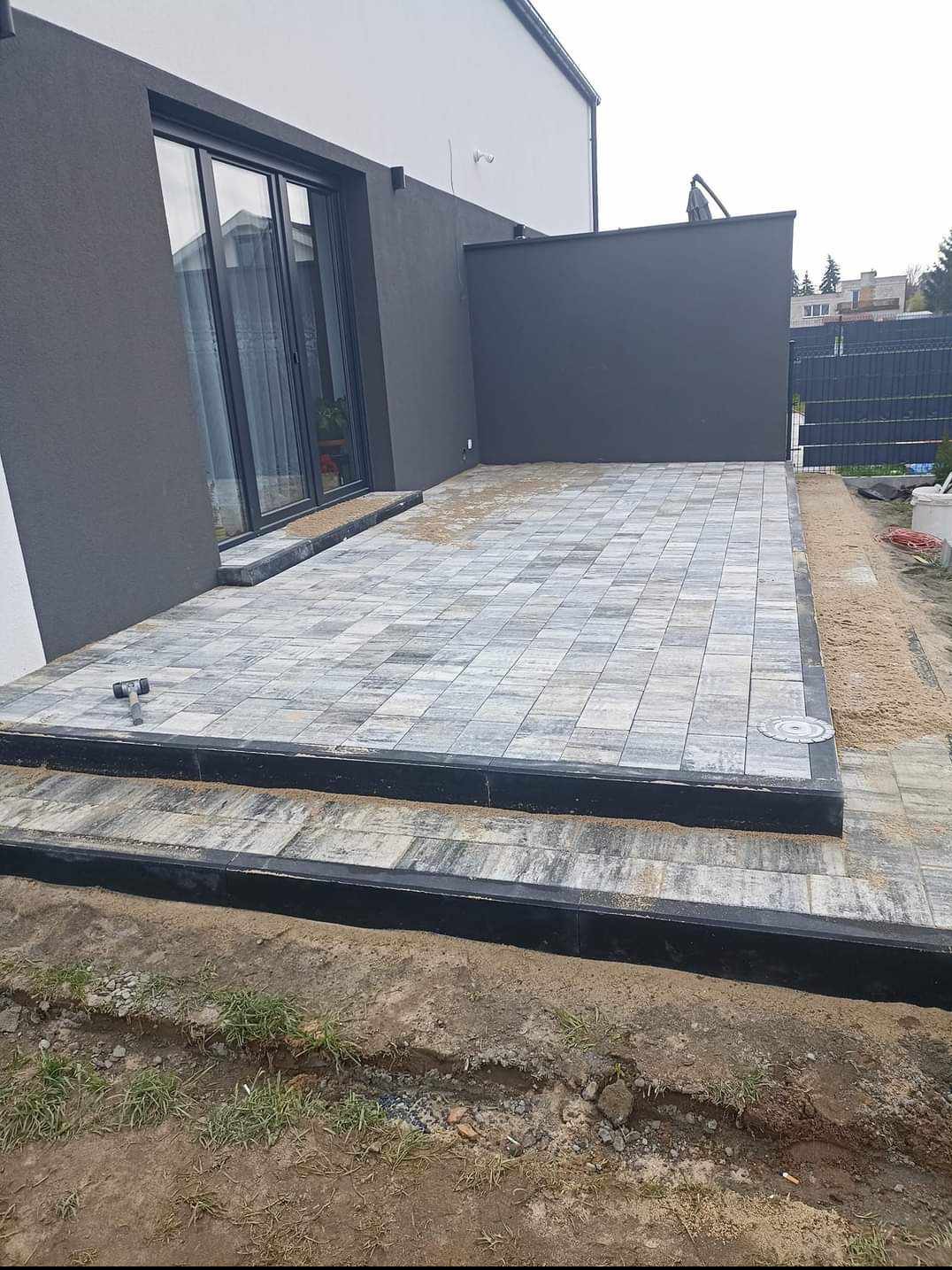 Nowoczesny taras z szarej kostki brukowej ułożonej w kwadratowy wzór, z czarnymi obrzeżami i dwoma stopniami przed wejściem do domu o szarej elewacji.