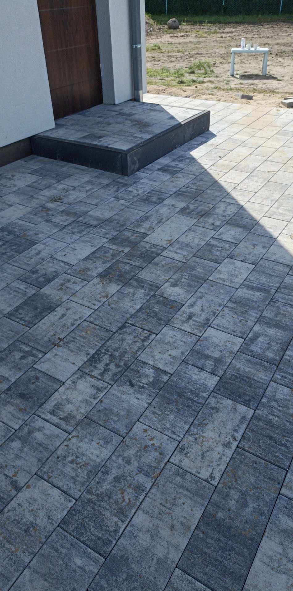 Ułożona kostka brukowa w odcieniach szarości prowadząca do schodów wejściowych, widoczny cień rzucany przez budynek.