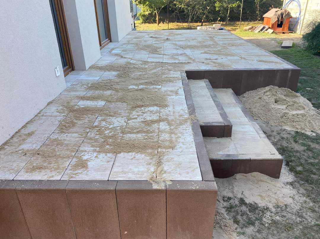 Etap prac: Jasnoszara kostka brukowa ułożona na tarasie i schodach, zasypana piaskiem do wypełnienia szczelin, widoczne brązowe obrzeża i fragment budynku z oknami w tle.
