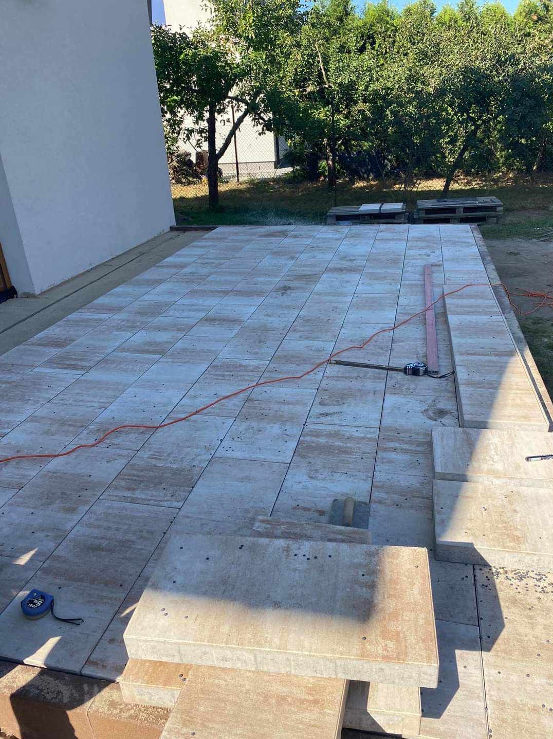 Wykończony taras z jasnych płyt z delikatnym wzorem, widoczne narzędzia i pomarańczowy kabel, w tle drzewa i fragment budynku.