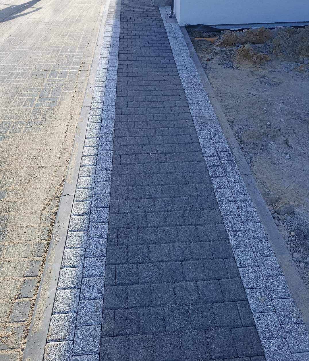 Ścieżka z ciemnej kostki brukowej obramowana jasną kostką granitową, prowadząca wzdłuż ściany budynku, z widocznym piaskiem i ziemią po bokach.