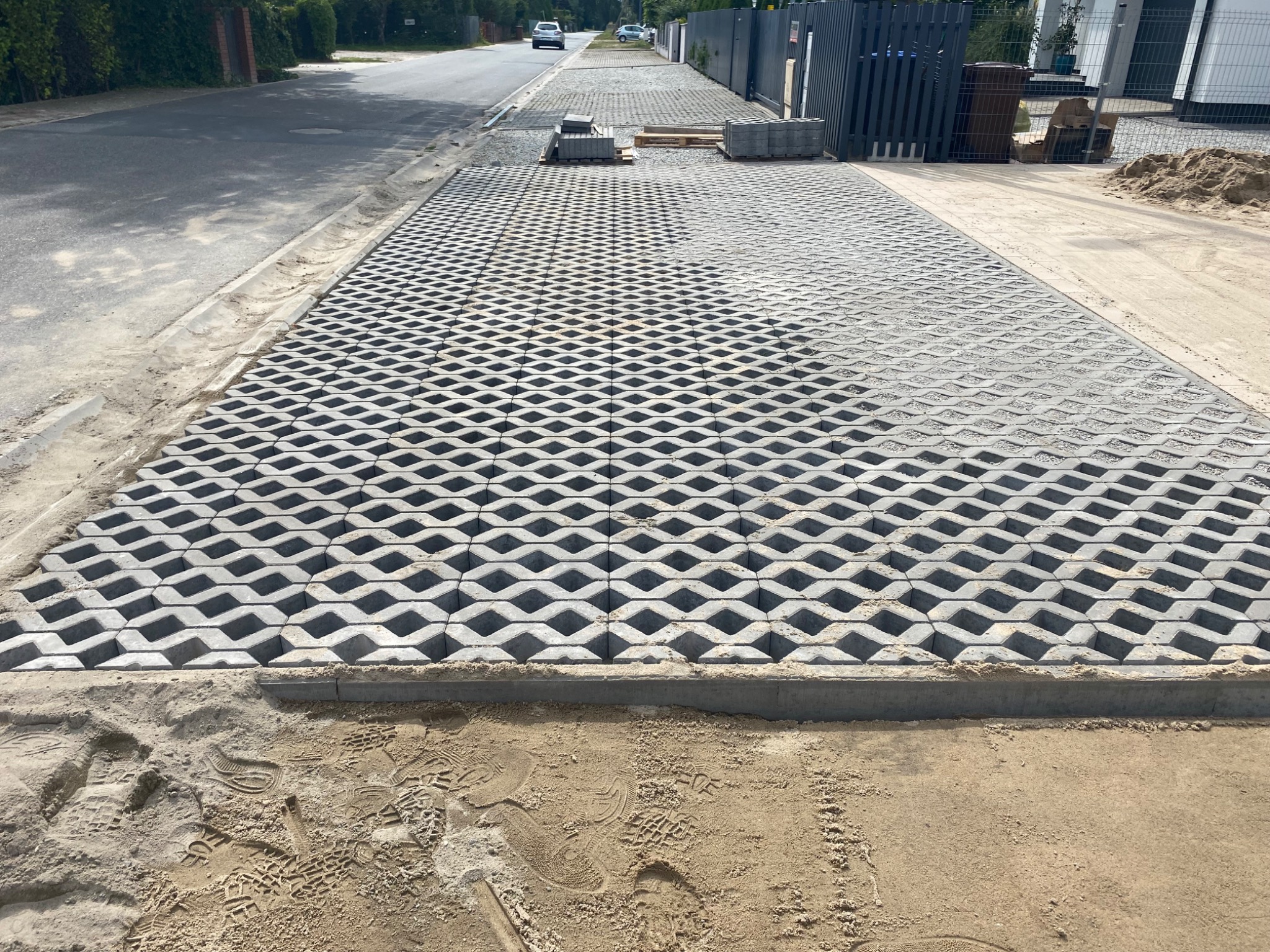 Widok z góry na ułożoną, szarą ażurową kostkę betonową na podjeździe, z widocznymi śladami stóp w piasku na obrzeżach i stosem kostki do ułożenia w tle.