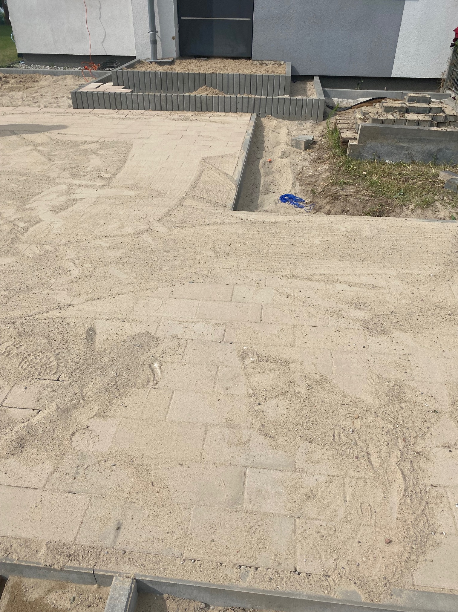 Niedokończona powierzchnia z kostki brukowej zasypana piaskiem, widoczne odciski butów, obrzeża z szarej kostki, fragment budynku w tle.
