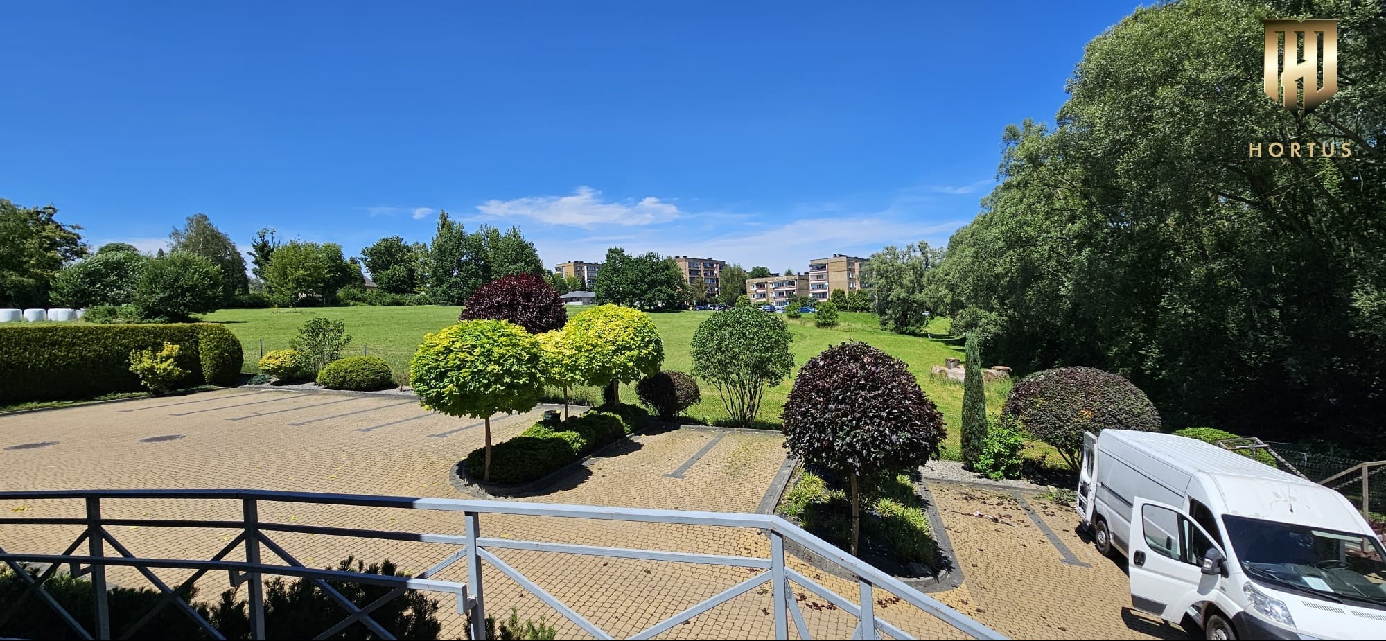 Widok na zadbany teren zielony z równo przyciętymi drzewami kulistymi, trawnikiem i budynkami w tle, widziany z góry zza metalowej balustrady, z logo firmy w prawym górnym rogu i białym samochodem...