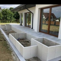 PROInvestment - Nowoczesny taras wyłożony szarą kostką brukową z przygotowanymi, betonowymi rabatami na rośliny wzdłuż ściany domu z dużymi, brązowymi oknami.