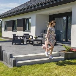 PROInvestment - Nowoczesny taras z szarej kostki brukowej, schody z jasnoszarego kamienia, niskie ogrodzenie z betonowych elementów, czarne meble ogrodowe, młoda kobieta schodząca po schodach w letniej sukience