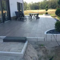 Nowoczesny taras wyłożony szarą kostką brukową, obrzeża z jasnego granitu, z elementami dekoracyjnymi w postaci kulistych krzewów i czarnych kamieni, widok na dom z dużymi oknami i ogród.